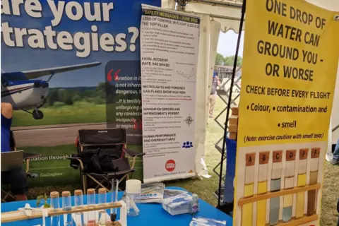 Air Accident Investigation Branch A display booth at an outdoor aviation safety event featuring tables with fuel sample test tubes, informational leaflets, and large posters. One poster warns that One drop of water can ground you – or worse, showing test tubes with different fuel contamination colours. Another banner discusses aviation safety messages. The setup is under a tent, with equipment and signage arranged on blue and yellow table coverings.