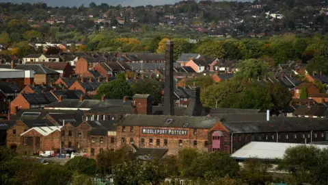 Getty Images Middleport Pottery seen from a distance, surrounded by other industrial buildings with houses and trees behind it extending up a hill. The brown brick building has a large Middleport Pottery sign in black and white high up. A chimney and kiln can be seen behind it.