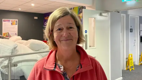 Fiona Bolt, who has blonde hair and is wearing a red jacket, is smiling at the camera. She is standing in front a pile of blankets.