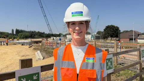 A woman in a hi-vis jacket stands in front of construction work. She is wearing a white hard helmet with the text Severn Trent at the front. Fencing and other equipment can be seen behind her as well as a large crane.