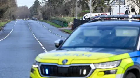 A police car, blurred, is parked across a main road. There are rows of trees and on either side of the road and a white building on the right. 
