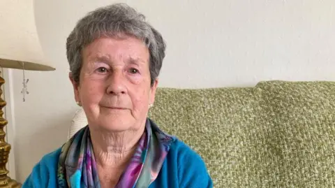 A woman with dark, greying hair, wearing a blue cardigan and a colourful scarf, sits on a couch with a green blanket hung over it. Half a lamp can be seen in the background. 