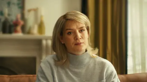 Netflix Blonde-haired woman wearing a grey turtleneck jumper sitting on the sofa looking at the camera 