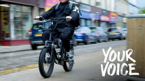 A man on a bike wearing all black. The background is slightly blurred with cars parked along the side of the pavement visible. On the bottom right hand corner is some text which says "YOUR VOICE".
