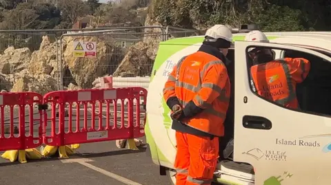 Two workmen in orange high-vis outfits stand near a van marked 'Island Roads' in the foreground. In the background, barriers and metal fences block off the road from the rockfall. 