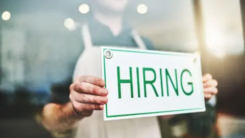 Person holding a sign with the word hiring in green letters on a white background.