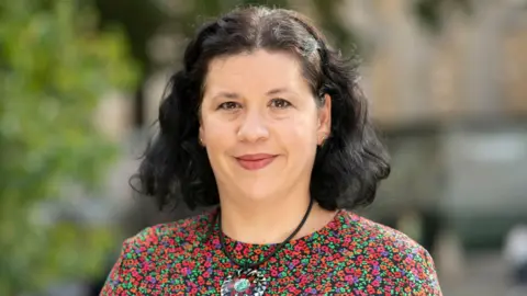 Silviya Barrett Silviya Barrett, who is wearing a bright floral top, has shoulder-length dark hair and is smiling, closed mouth.