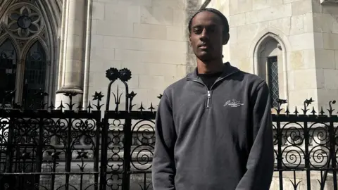 Champion News Man in his 20s wearing a grey hoodie stood outside of the Central London County Court black railings behind him, and the grey stone building.