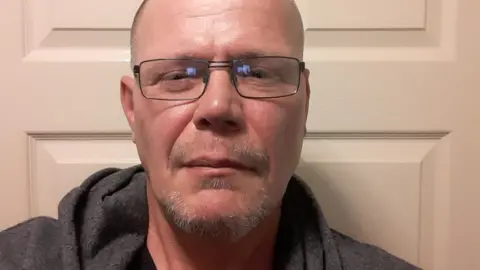 Stuart Vale, a man with a bald head and glasses is wearing a grey hooded top. He is standing against a cream-coloured door.