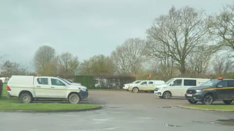 Supplied A car park positioned in front of trees which is filled with vans and trucks. 