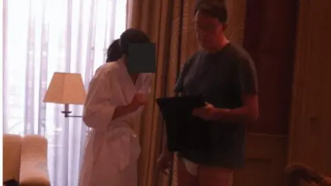 US Department of Justice Mandelson is seen standing next to a woman in a white robe. Her face is redacted. He is wearing a blue T-shirt, and white underwear. He is holding what appears to be an i-Pad.