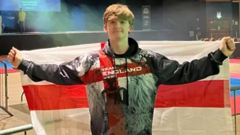 Family handout/PA Alex Eastwood in a Team England jacket holding an England flag behind him