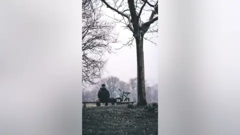 Yu Wang The back of a young man, alone on a park bench, with leafless winter branches framing him in the image. It is a portrait photo. One straight, thin tree strands to the right of the frame.