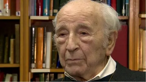 Chaim Ferster, with thinning white hair, speaks into the camera with a serious expression in a room surrounded by bookshelves. 