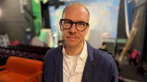 Derek Bond is standing in the middle of the picture with a blue jacket and white shirt - he has glasses and in the background is stage with theatrical props and furntiture.