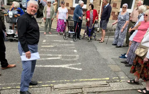 Rev Sheila Bawden standing partly on the road and kerb in front of wooden sleeper. She is surrounded by people, some with mobility scooters, walking frames and walking sticks. 