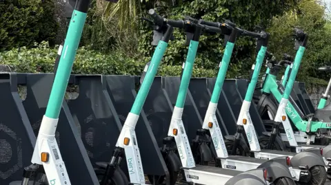 A row of white and mint green Beryl e-scooters parked up in a rack on a street