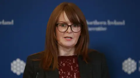 Caoimhe Archibald wearing a dark suit and red shirt. Behind her is a blue Northern Ireland executive board. 