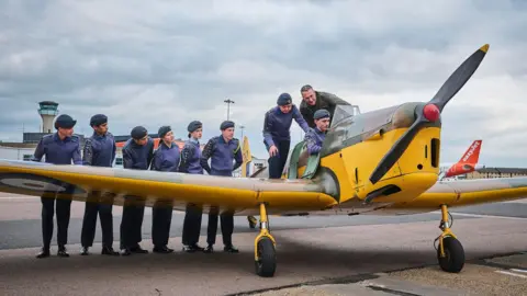 The Miles Magister yellow plane on the forecourt at Luton Airport. A group of eight cadets wearing their purple and black uniform and caps stand behind the left wing, with one sitting in the cockpit and two standing behind him. The pilot wears a green uniform and stands behind the cockpit. The airport control tower is in the background and you can see the orange tail of an easyJet plane.