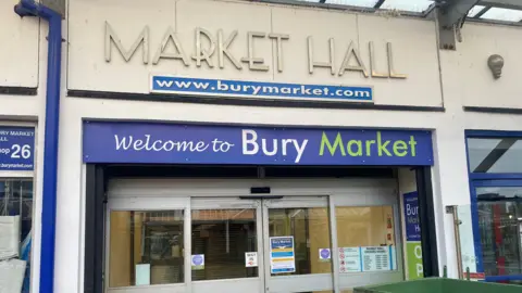 The entrance to the indoor section of Bury Market, which consists of glass sliding doors above a blue sign with the words 'Welcome to Bury Market'.