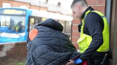 A police officer holding an arrested person from behind while walking away from a building. The person arrested has a coat over their head.