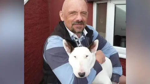 Family handout A man with a bald head and goatee beard looking at the camera and holding a white dog with black ears.