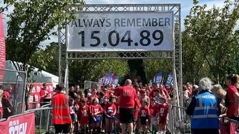Erica Dillon Dozens of children in red tops stand at the start line under a sign saying "Always remember 15.04.89". Spectators watch from the sides.
