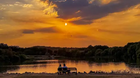 BBC Weather Watchers/ Raj's Range A couple with their backs to the camera observe the eclipse from a park bench, overlooking a lake