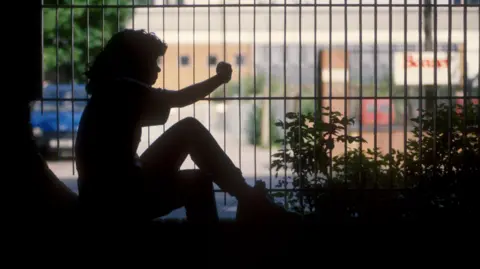 BBC A silhouette figure of a young girl sitting behind some railings.