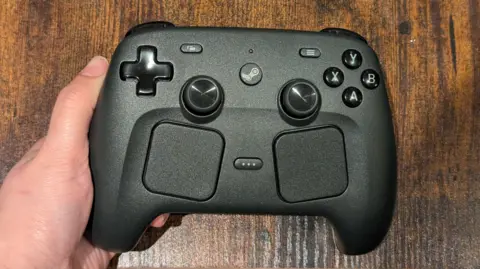 The Steam Controller, a gamepad, in charcoal black, with a four-way direction button, two thumbsticks, two trackpads, and various other buttons, held in a person's hand