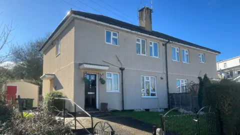 A property on Cobhorn Drive in Bristol. The flat is beige in colour with several windows on the front. The front door is black and to the side. There are hanging baskets either side of the door.