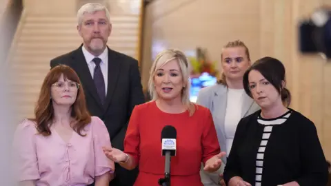PA Media (left to right) Economy Minister Caoimhe Archibald, Finance Minister John O'Dowd, Stormont First Minister Michelle O'Neill, Junior Minister (Executive Office) Aisling Reilly and Infrastructure Minister Liz Kimmins during a press conference in the Great Hall in Stormont, Belfast. O'Neill is speaking into a microphone. She has blonde shoulder-length hair and is wearing a red dress.