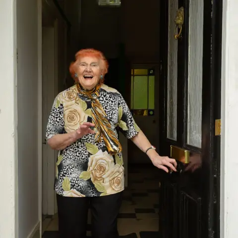 Maraya Gibbs Portrait of an elderly lady with red hair standing in her doorway