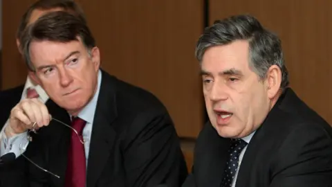 Getty Images Peter Mandelson holds his glasses in his hand as he looks at Gordon Brown, who is speaking. Both wear dark suits.