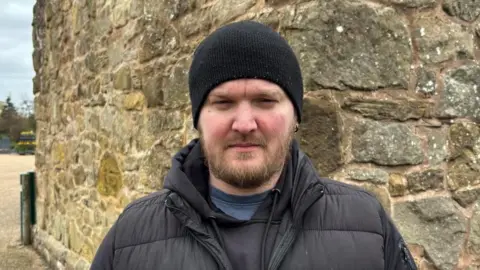 There is a man in a black beanie hat, and a brown beard. He is wearing a black puffer coat with a black hoodie and a blue t shirt. He is standing in front of a cobbled wall.