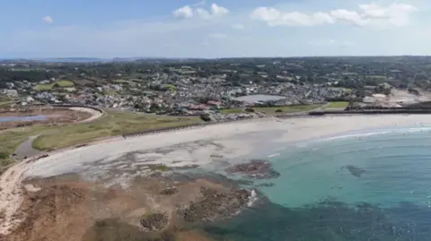 BBC A sandy beach with blue coloured sea and rocks on left and housing behind with some areas of greenery.