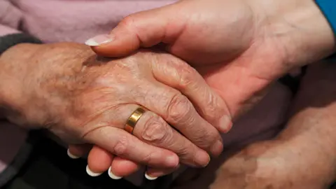 Close up image of the hand of an elderly person - wearing a gold ring on one finger - being held by a the hand of a younger person with manicured nails.