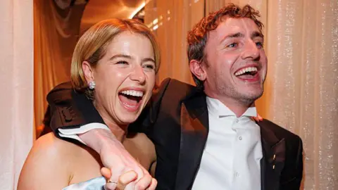 Getty Images Buckley with short blonde hair, diamond earring and off the should dress is smiling and she walks along with Paul Mescal. His arm is around her shoulder as he smiles. He is wearing a black suit jacket, white shirt and has short dark hair. 