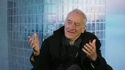 BBC Robert De Niro, sitting in front of a blue background, wears a black coat and a dark scarf. He has short grey hair and is gesturing with both hands out in front of him. 