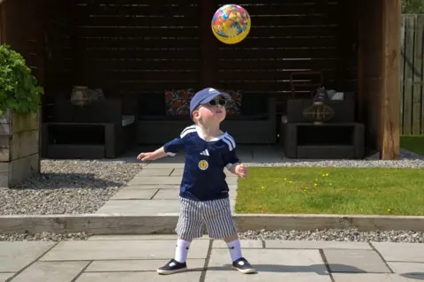 Kirsty Maclean A young boy wearing a Scotland football top and blue and white striped knee-length shorts, white socks and blue sandals, as well as sunglasses and a baseball cap, in a garden. A bright ball is mid-air and he is looking at it.
