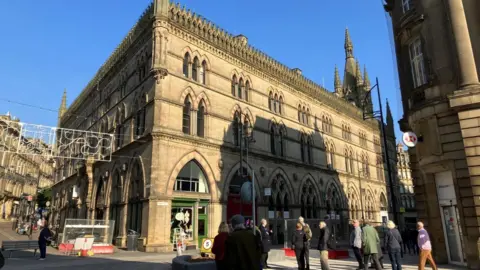 James Hughes Bradford's Wool Exchange building, with pedestrians stopping to admire the building. Barriers and fencing block access to areas currently under construction during paving work. 