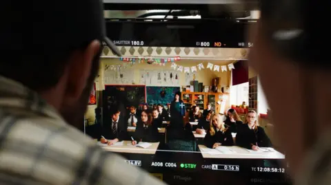 Laura Guest Shot of children in a classroom being viewed through the camera lens so you can see people's shoulders in the foreground. 