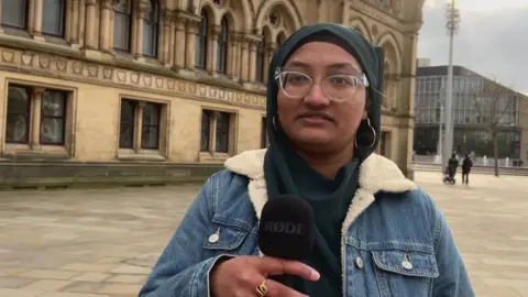 Aisha Iqbal/BBC A young woman wearing a blue headscarf and denim jacket. She is standing in front of a historic building in a city square. She wears glasses. She is holding a microphone.