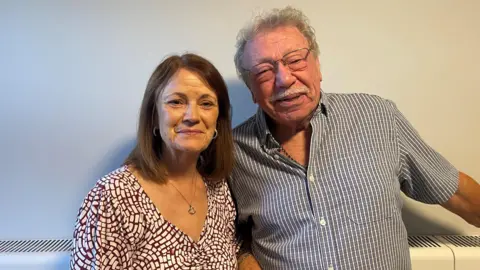 A woman with long brown hair and a white and brown dress, stands next to a man with short grey hair, glasses, a moustache and a grey and white shirt.