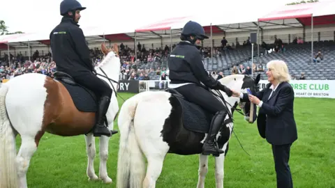 Eddie Mulholland/PA Wire Queen Camilla is standing by two horses - one white and brown and the other white and black - with riders dressed all in black - they have their backs to the cameras. The Queen is wearing a blue trouser suit and is shaking the hand of one of the riders and handing over a blue rosette. There are spectator stands in the background