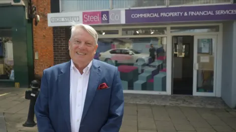 Jamie Niblock/BBC A man with grey hair wearing a light blue suit with a white shirt smiles at the camera. He is standing front of a High Street shop.