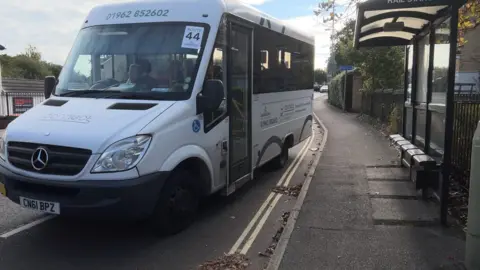 The number 44 bus at a bus stop. It is a white mini bus type and the bus stop is on the right hand side.