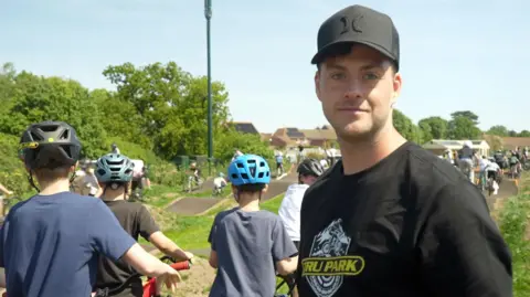 Connor Bennett/BBC Jono Jones is looking at the camera wearing a black T-shirt and baseball cap. Children with bicycles can be seen behind him.