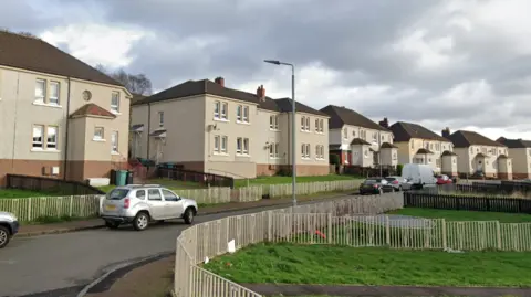 Google A residential street with beige buildings with dark rooves. They have fenced off front gardens and cars are parked along the street.