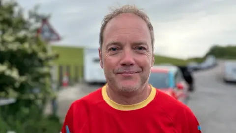 Warren Porritt in a red t-shirt, looking at the camera. He is smiling slightly but appears tired. A road with parked cars is blurry but visible in the background.